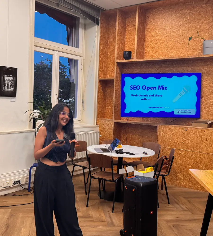 A woman holding the microphone in front of a screen that reads "SEO Open Mic, grab the mic and share with us", in a room with wooden shelves.