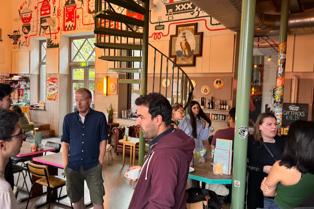 People standing and talking with drinks at Brouwerij Poesiat & Kater, by a green spiral staircase and murals on the walls.