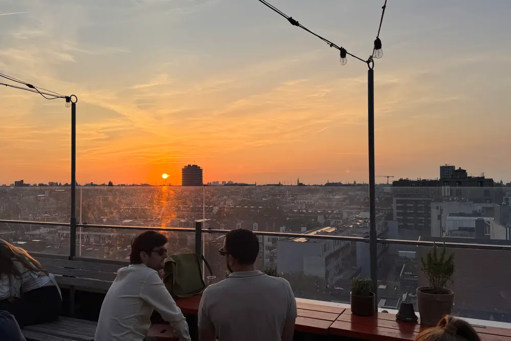 The sun going down over Amsterdam from the Canvas rooftop, with people sitting along the railing under string lights.