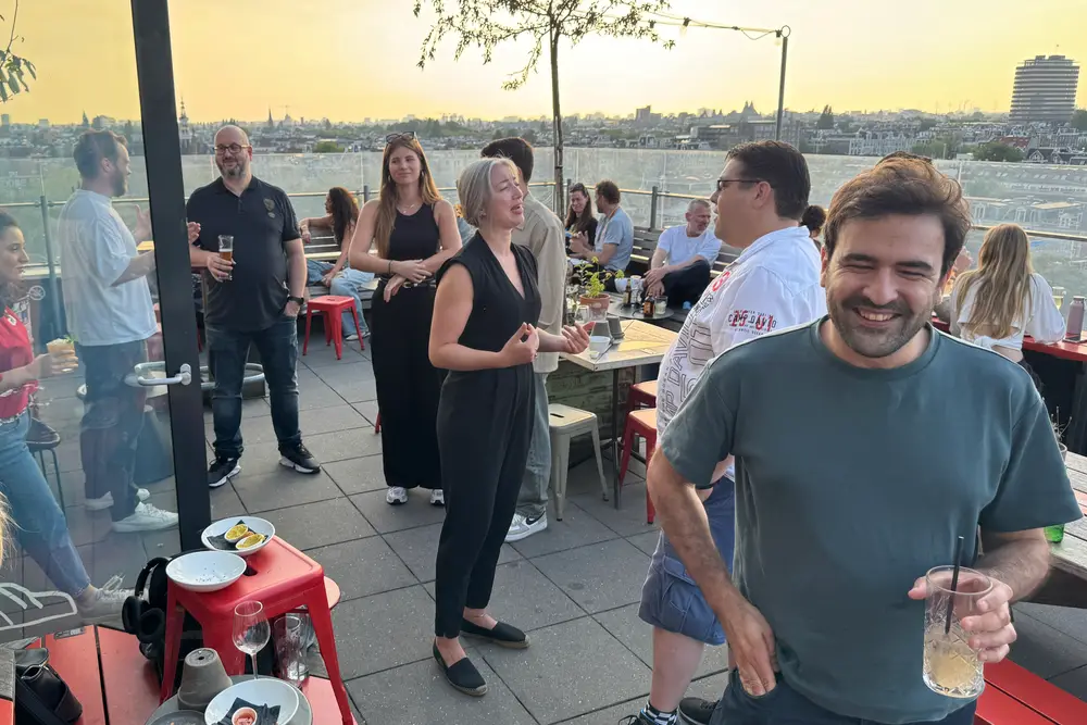 People with drinks on the Canvas rooftop terrace in the evening sun, laughing and talking, with the Amsterdam skyline behind them.